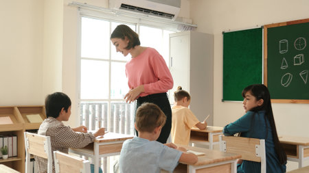 Caucasian teacher walking and check student homework at classroom while diverse children doing classwork at classroom. Diverse smart student lecturing and taking a note at elementary school. Pedagogy.の写真素材