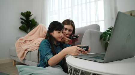 Happy girl playing game and sitting while mom lie on sofa by using laptop screen. Caucasian parent and attractive girl spend time together while holding joystick and focus on winning games. Pedagogy.の写真素材