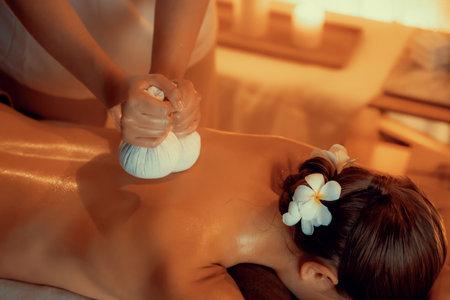 Hot herbal ball spa massage body treatment, masseur gently compresses herb bag on woman body. Tranquil and serenity of aromatherapy recreation in warm lighting of candles at spa salon. Quiescentの写真素材