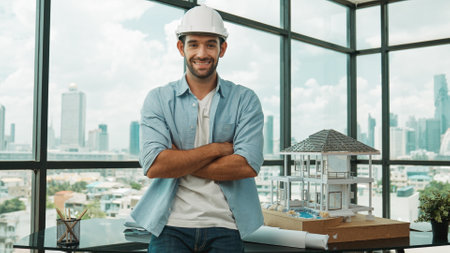 Portrait of architect engineer in casual outfit smile at camera while crossing arms. Engineer looking at camera and sitting with arms folded near house model while wearing safety helmet. Traceryの写真素材