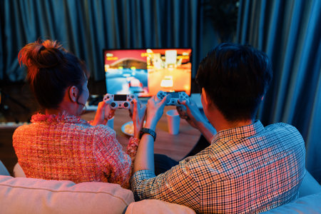Couple gamer with controlled joystick playing car racing video game together on tv screen with snack at back side view, sitting sofa at neon light colorful living room at modern comfy home. Infobahn.の写真素材