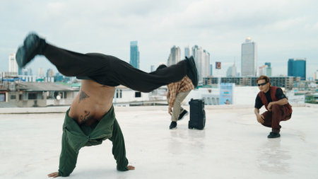 Multicultural hipster practice B-boy dance at rooftop with city view. Professional break dancer doing freeze pose while diverse hipster looking or encouraging his friend. Outdoor sport 2024. Hiphop.の写真素材