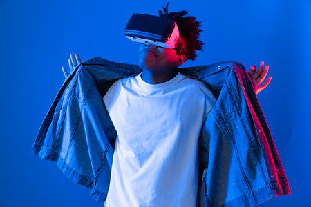 Smiling young African American looking through VR dancing in song with hologram gaming on pink blue...