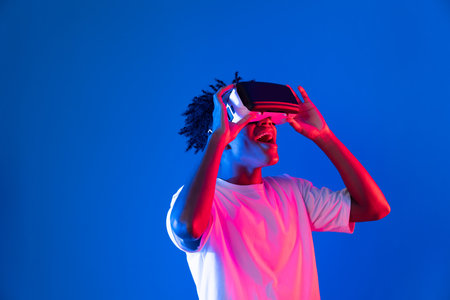 Surprised young African American looking through VR holding interesting object hologram on pink blue neon wall at metaverse world connecting digital futuristic technology virtual reality. Contrivance.の写真素材