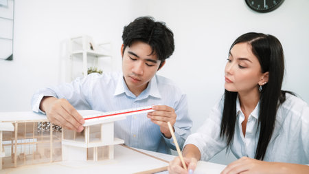 Professional male engineer measure house model by using ruler while beautiful cooperative coworker working together by writing on blueprint. Creative design and teamwork. Closeup. Immaculate.の写真素材