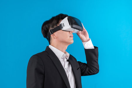 Smiling Asian businessman looking thorough VR connecting to business world report in futuristic metaverse, analyzing innovation technology ads in virtual reality isolated blue copyspace. Contrivance.の写真素材