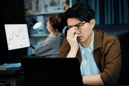 Businessman with stressful face working on laptop waiting email while coworker trading stock...