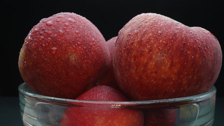 Close-up showcases a crisp bright red fresh apple nestled comfortably in a glass bowl. The black...
