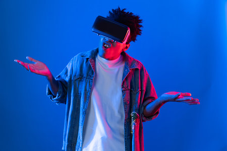 Young African American wearing VR pointing to empty space product launching brand explaining detail on blue pink neon light wall futuristic technology virtual reality at metaverse world. Contrivance.の写真素材