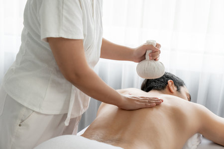Hot herbal ball spa massage body treatment, masseur gently compresses herb bag on man body. Tranquil and serenity of aromatherapy recreation in day lighting ambient at spa salon. Quiescentの写真素材