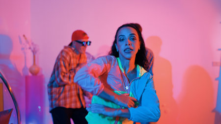 Attractive happy dancer looking at camera while smart woman moving at rhyme with neon light. Professional hispanic performer break dancing while wearing colorful cloth with diverse friend. Regalement.の写真素材