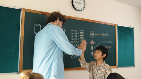Attractive boy writing answer while teacher give high five to encourage child. Attractive student walking back to his seat while teacher applause and clap hand to schoolboy at Math lesson. Pedagogy.の写真素材