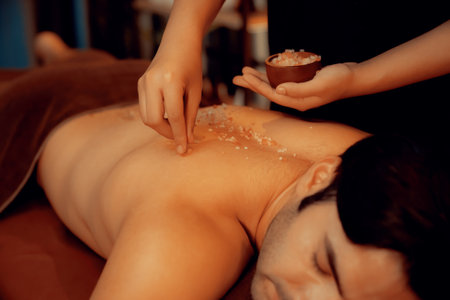 Man customer having exfoliation treatment in luxury spa salon with warmth candle light ambient. Salt scrub beauty treatment in Health spa body scrub. Quiescentの写真素材