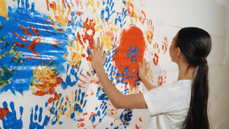Asian girl use hand to stamp the wall with red color in art lesson. Multicultural highschool student...
