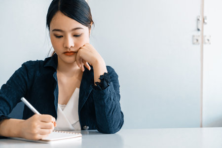 Beautiful young Asian woman writes letter on notebook while sitting at office desk. Content writer and secretary job concept. udsの写真素材