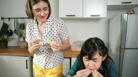 Energetic caucasian mother and asian girl cooking together and eating orange or preparing salad for dinner. Happy mom and daughter making healthy food with fresh food. Healthy food concept. Pedagogy.の写真素材