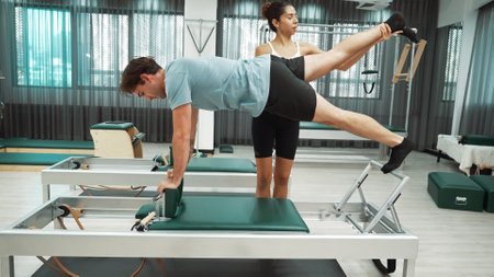 Skilled Pilates instructor assists a client in performing a plank with a leg extension on a reformer machine. Female trainer assist side plank variation on reformer for improving balance. Habituate.の写真素材