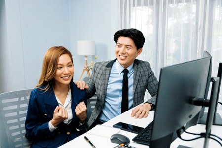 Successful smiling Asian business partners raising fist up together in profitable project job in stock exchange database on pc wearing formal suit at modern home office at side view image. Infobahn.の写真素材