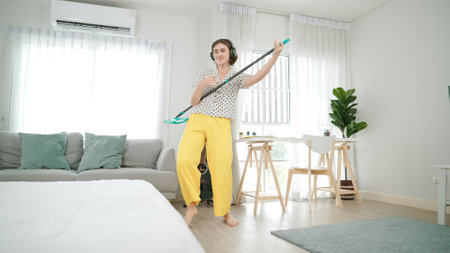 Smiling housekeeper with headphone listen music while cleaning floor. Happy caucasian mother mopping floor while dancing to lively song at living room. smiling mom doing housework. Lifestyle. Pedagogyの写真素材