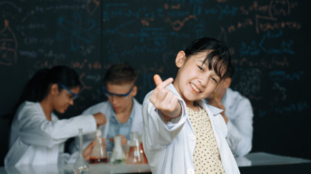 Asian scientist show miniheart to camera while diverse group doing experiment. Young schoolgirl standing at blackboard with chemical theory written while wearing lab coat at laboratory. Edification.の写真素材