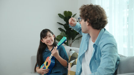 Back view of happy father looking at smart daughter playing ukulele warmly and singing song. Happy dad motivated and encouraged american school girl enjoy playing guitar. Family recreation. Pedagogy.の写真素材