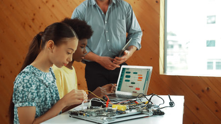 Teacher standing while explain coding and robotic construction. Diverse teenager fixing mother board and using electronic tool while looking at laptop display programing code screen. Edification.の写真素材