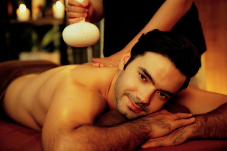 Hot herbal ball spa massage body treatment, masseur gently compresses herb bag on man body. Tranquil and serenity of aromatherapy recreation in warm lighting of candles at spa salon. Quiescentの写真素材