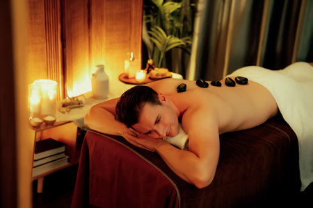 Hot stone massage at spa salon in luxury resort with warm candle light, blissful man customer enjoying spa basalt stone massage glide over body with soothing warmth. Quiescentの写真素材