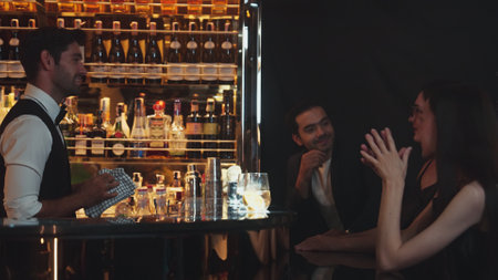 Smart bartender serving gin fizz or tonic with sliced lemon at luxurious bar, engaging lively conversation with VIP customer couple at side view in atmosphere at lighting warm nightclub. Vinosity.の写真素材