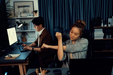 Businesswoman working on desk with stretching arm up and down manner with body health ache of strain overwork while coworker trading stock market on website on pc at late over night time. Infobahn.の写真素材