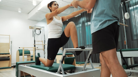 Woman receiving assistance in split stretch exercise during pilates class on trapeze table for flexibility and core training. Student doing split stretch on reformer for improving balance. Habituate.の写真素材