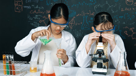 Young highschool student doing experiment by pouring sample in test tube. Girl wearing glasses and looking under microscope to inspect sample in STEM science class. Creative education. Edification.の写真素材