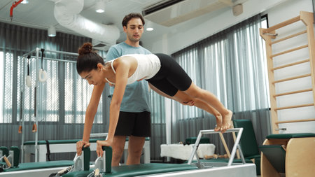A Pilates instructor guide a client through a plank variation on a reformer machine. Healthy woman doing side plank variation on the reformer with trainer assistance for improving balance. Habituate.の写真素材