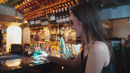 Beautiful woman guest zipping to taste in Christmas blue Lagoon cocktail with good mood sitting on luxurious counter bar green neon snow Christmas pine tree festive theme on wine background. Vinosity.の写真素材