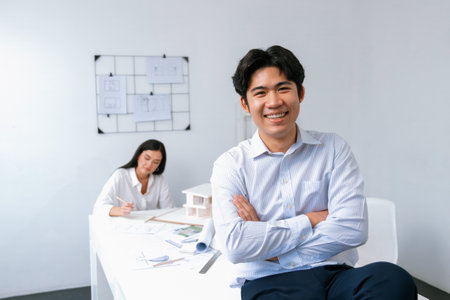 Professional asian businessman crosses arms with confident while young beautiful caucasian colleague drawing a blueprint on meeting table with house model and blueprint at modern office. Immaculate.の写真素材