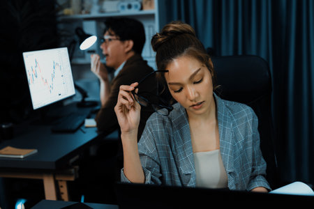 Stressful businesswoman looking at computer stock exchange via email with coworker working at back position talking to ask problem with loss profit on time at modern home office at night. Infobahn.の写真素材