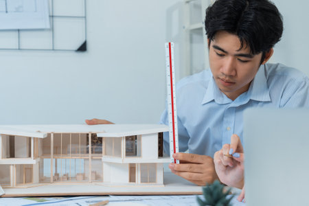 Professional male asian architect using ruler to measure house model length while young beautiful caucasian colleague using laptop to analyzed data on meeting table with house model. Immaculate.の写真素材