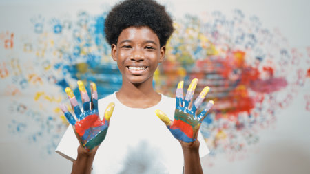 African boy looking at camera while showing two side of hand to camera with colorful color. Smiling happy highschool teenager wearing white shirt while standing at colorful stained wall. Edification.の写真素材
