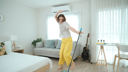 Caucasian housekeeper mopping the floor while dancing in lively mood at modern apartment. Happy energetic mother or cleaner enjoy cleaning the living room while moving to music. Lifestyle. Pedagogy.の写真素材