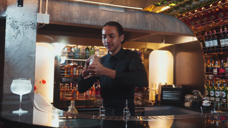 Asian smart bartender shaking mixed cocktail preparing to serve at luxurious counter preparation,...