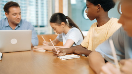 Young smart teenager writing and talking about topic at classroom. Diverse highschool student taking a note in other notebook while sitting with multicultural friends. Creative education. Edification.の写真素材