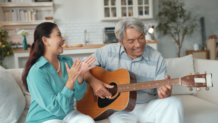 Happy senior man sitting at sofa and playing guitar or music instrument while his wife singing and moving to music. Elder couple spending time together and enjoy doing activity together. Myrmidon.の写真素材