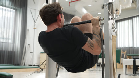 Client performing an inversion exercise on the Pilates Cadillac with instructor guidance. Attractive man hanging at bar for building upper body strength and increasing core and stability. Habituate.の写真素材