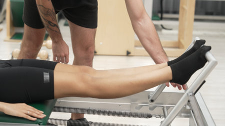 Close up of trainer arranging woman posture while student using reformer at gym. Pilates instructor providing support to woman during leg strengthen exercise using pilates reformer machine. Habituate.の写真素材