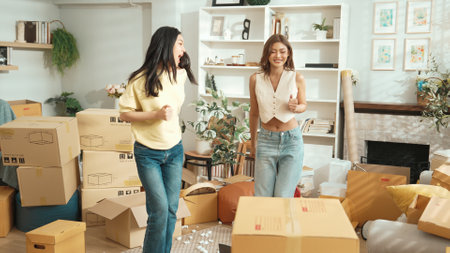 Happy lesbian lover dancing together at living room in moving day. Couple of friend moving to music and perform freestyle dancing at living room with cardboard box scattering. Relocation. Myrmidon.の写真素材