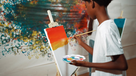 Closeup of african boy hand paint canvas with warm tone color while caucasian girl in casual outfit drawing cool tone colors behind at colorful stained wall. Creative activity concept. Edification.の写真素材