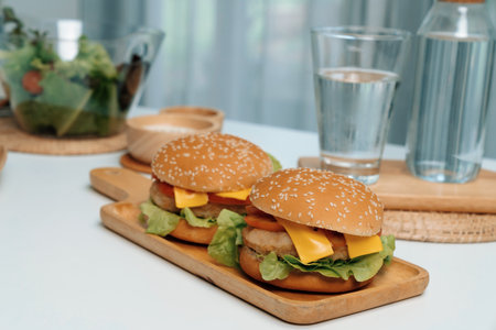 Focusing on double giant fried meat hamburger meal on square handle wooden board. Decorated by salad, homemade bun bread, beside drinking water on fast healthy food concept on white table. Infobahn.の写真素材