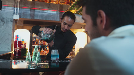 Bartender pouring mixed blue lagoon cocktail liqueur to cooling glass while guest talking friends...