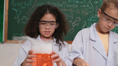 Closeup of students in lab coats and safety glasses examining a solution during a science experiment. A chalkboard with chemical formulas can be seen in the background while they conduct their discovery work.の写真素材