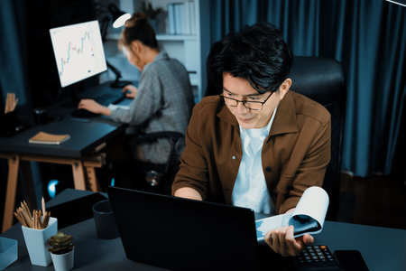 Businessman with stressful face looking on document file deadline while coworker trading stock exchange investment at late night time at modern office working on startup company project job. Infobahn.の写真素材